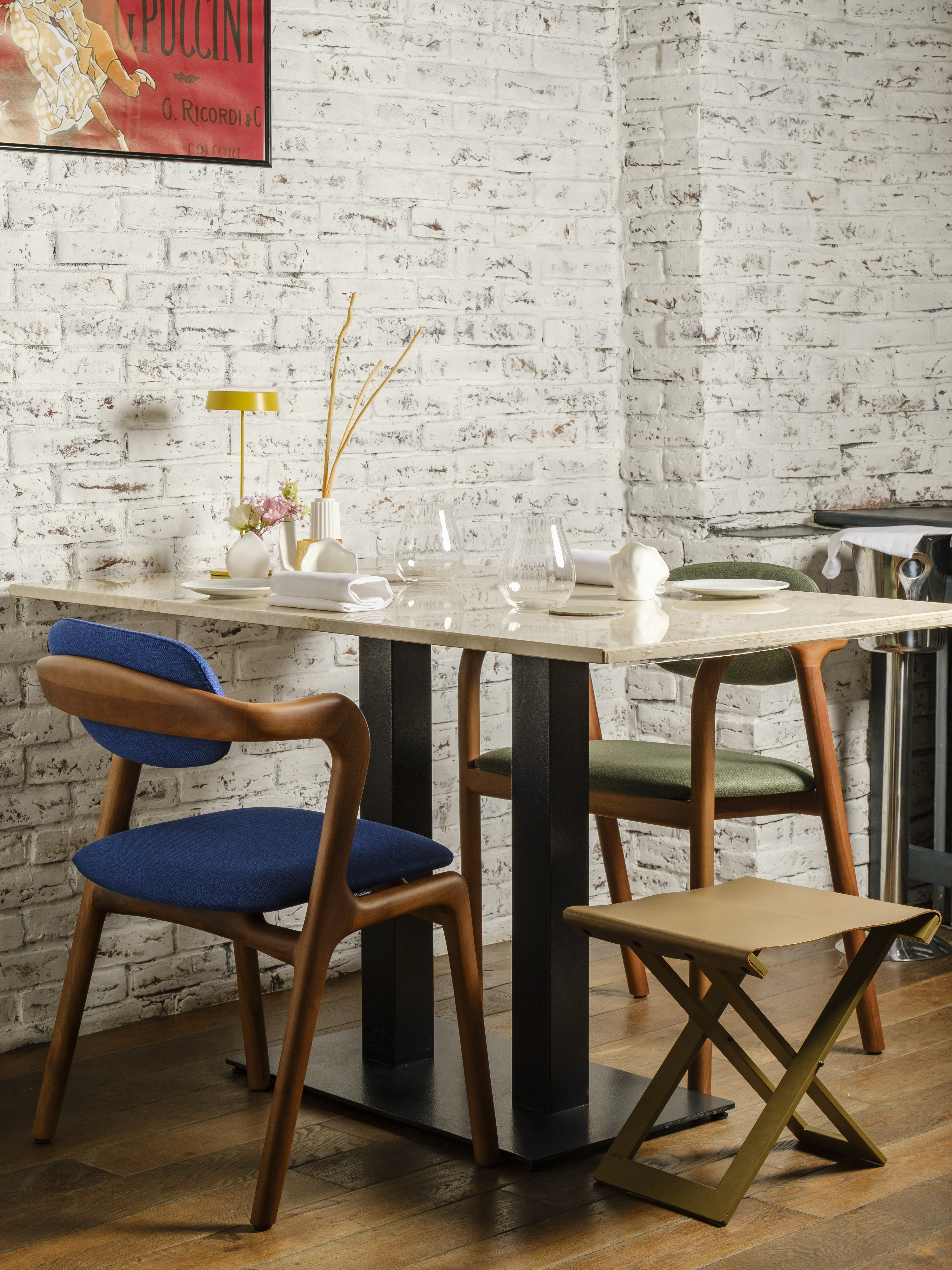 A quiet corner table against white brick, two blue chairs, a brass lamp and a folded napkin