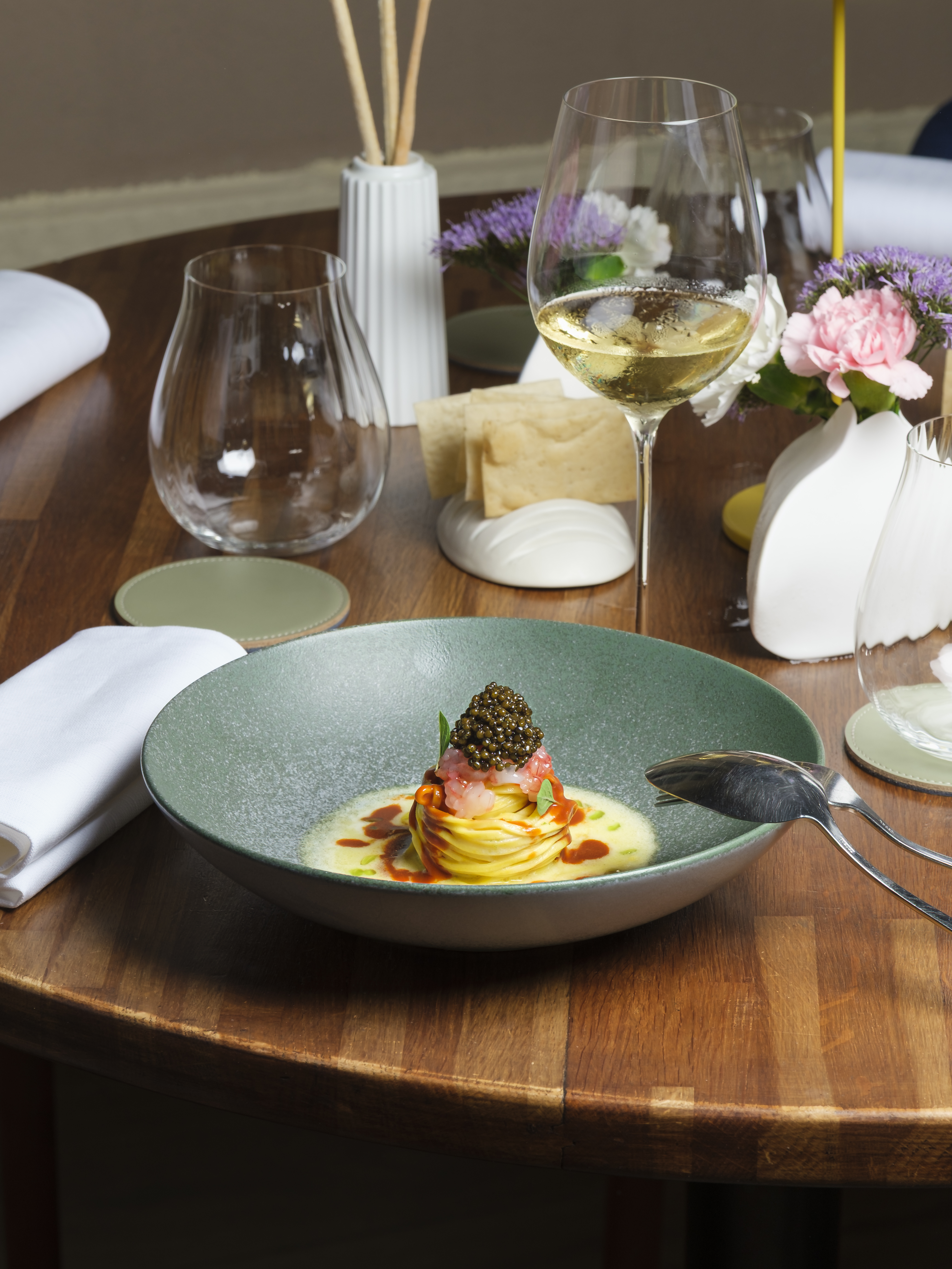 Tagliolini with gambero rosso and caviar in a green bowl on a walnut table, glasses and flowers behind