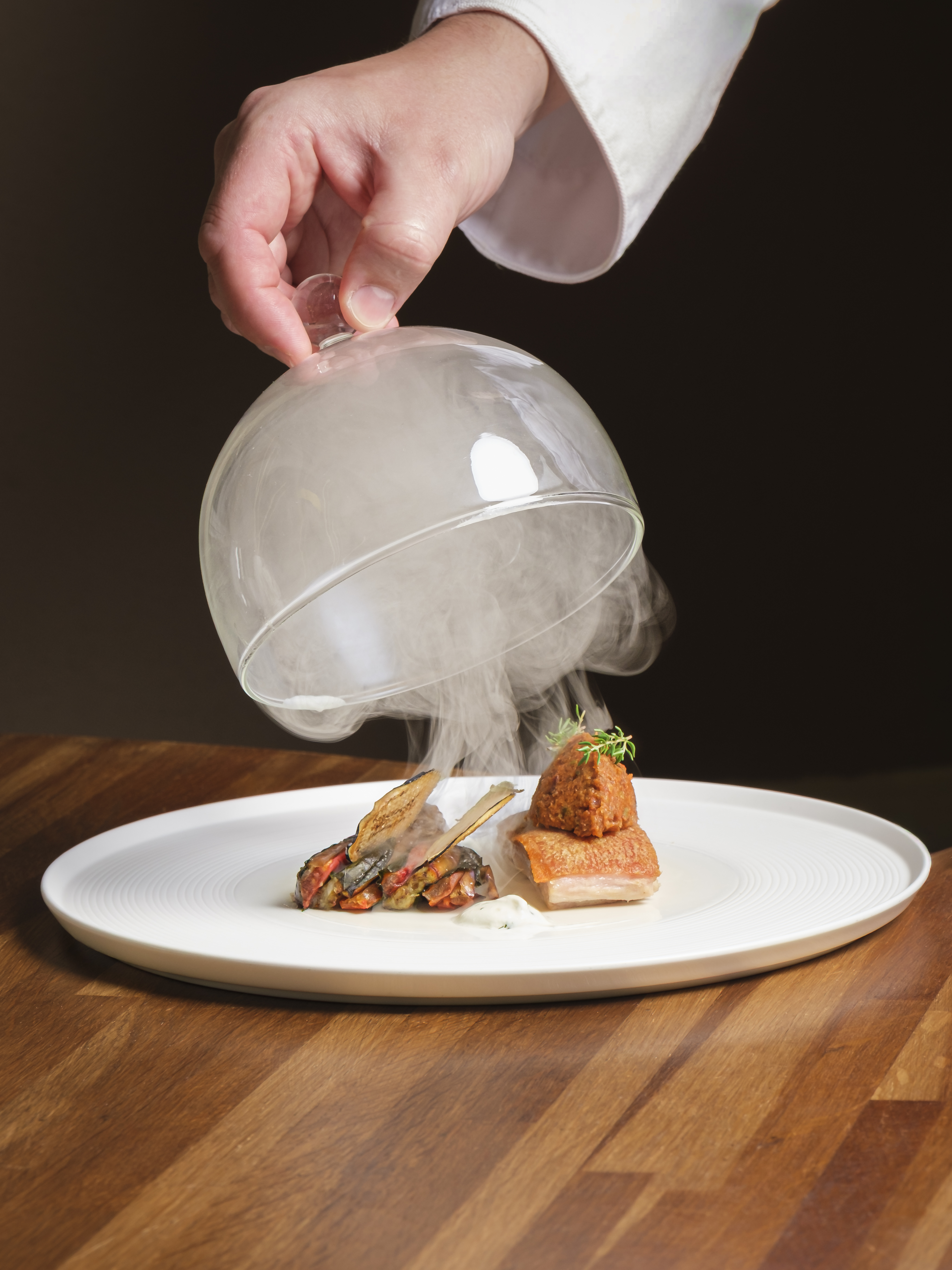 A glass cloche lifted from a plate, smoke curling out over the dish