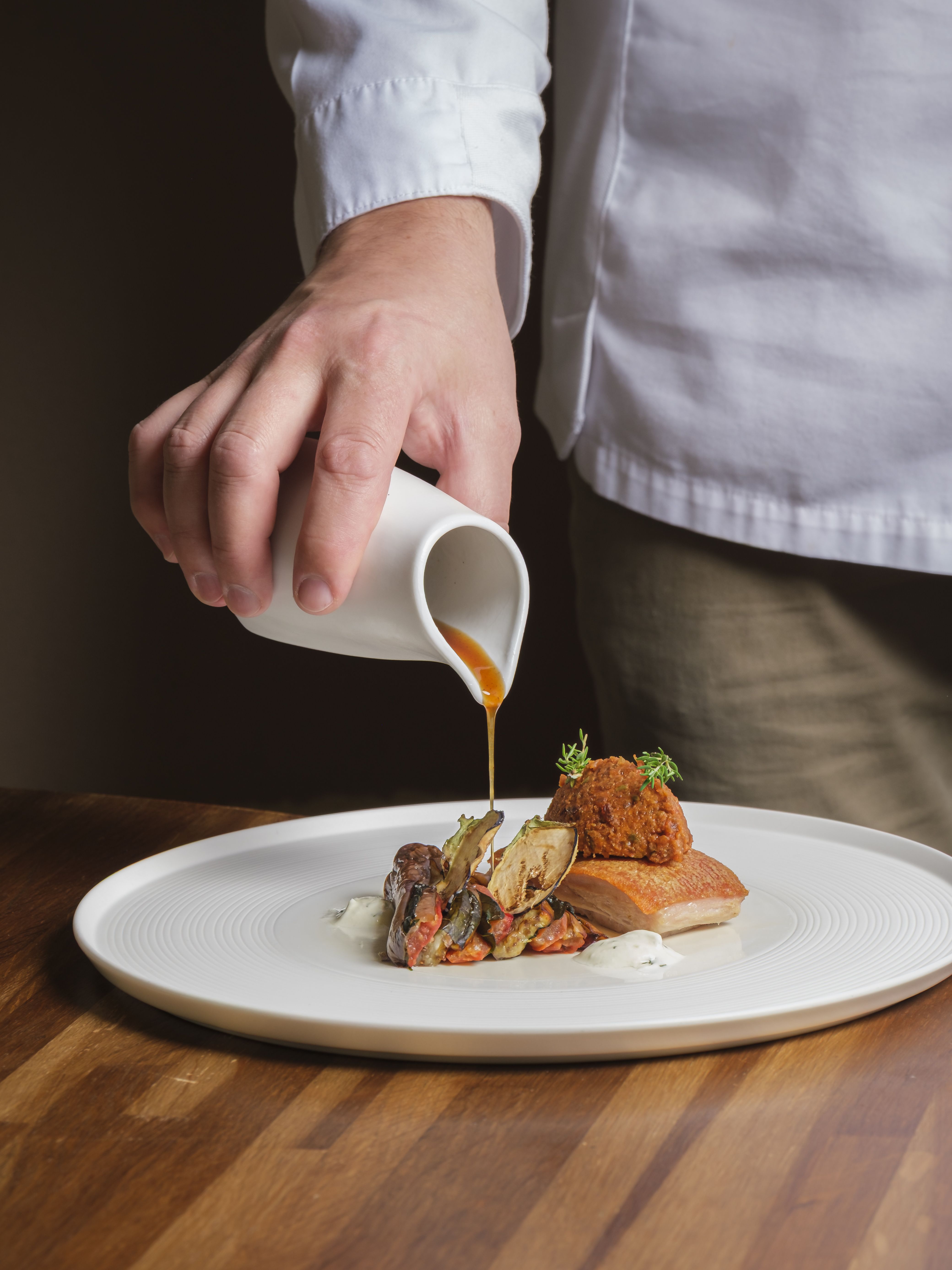 The chef pouring a jus from a small jug over a plated main course