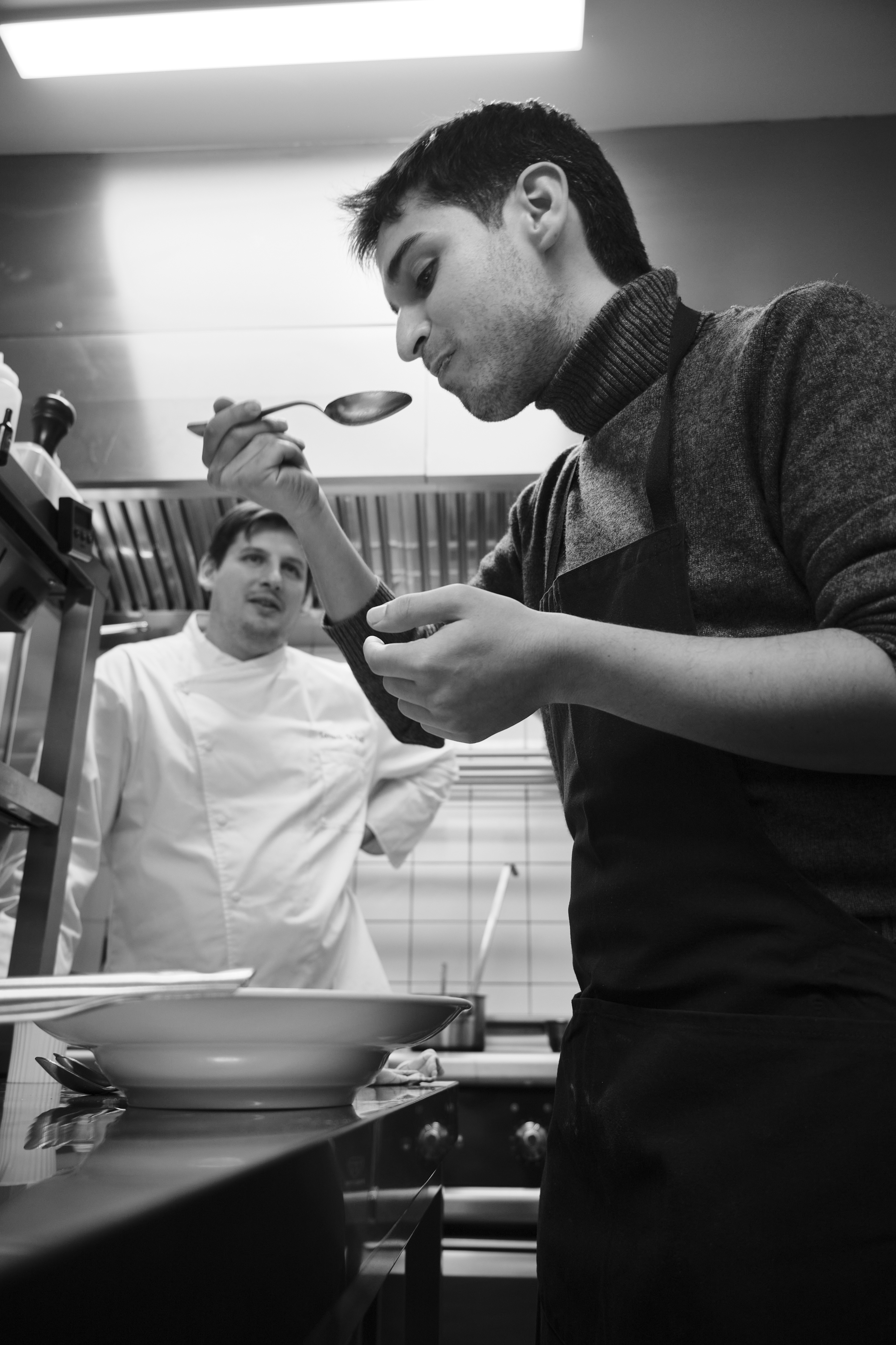 A cook tasting from a spoon over the pass while the kitchen works, in black and white